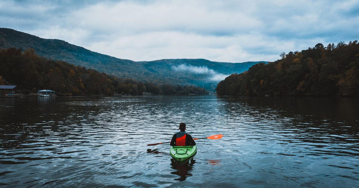 A kayaker paddling alone across a calm lake below forested hills