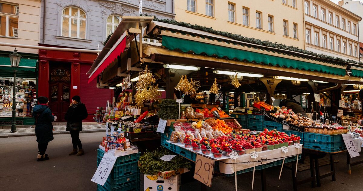 An open-air market square with produce stalls and shoppers walking between them
