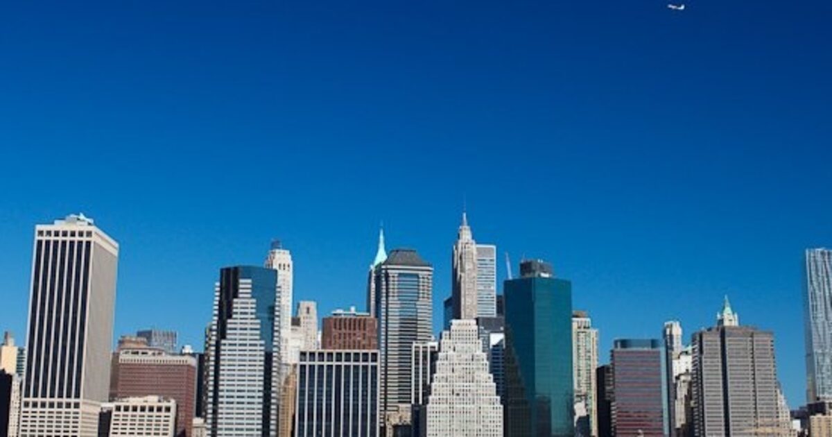 A dense Manhattan skyline of skyscrapers built over decades, with a single small plane passing overhead