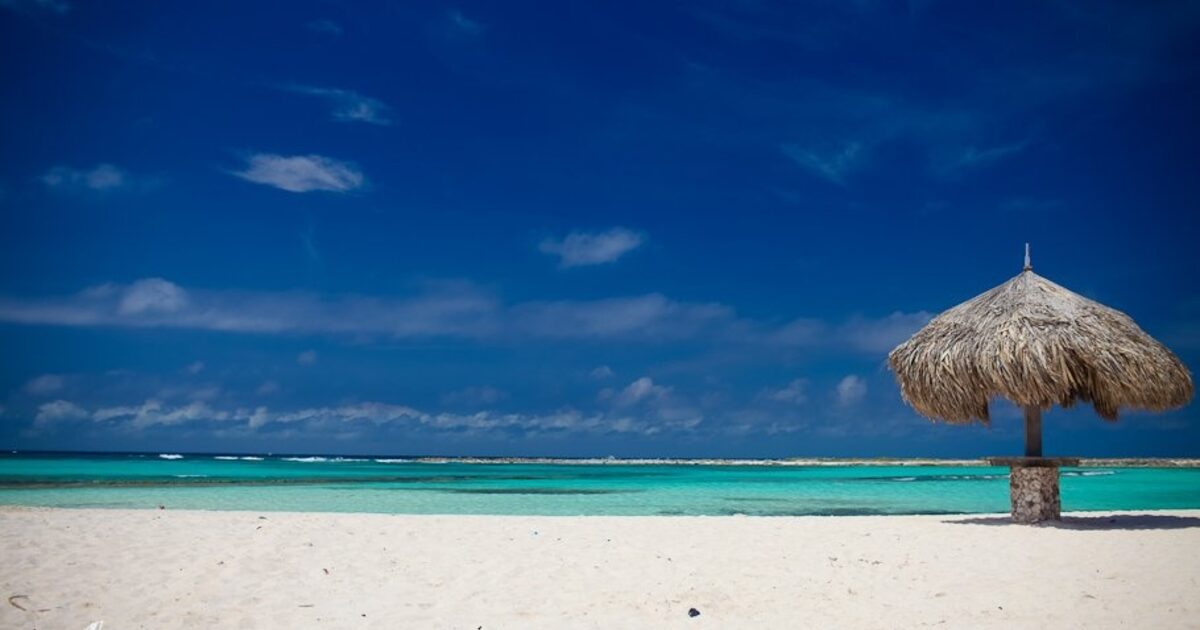 An empty white-sand beach with a single thatched palapa and turquoise water
